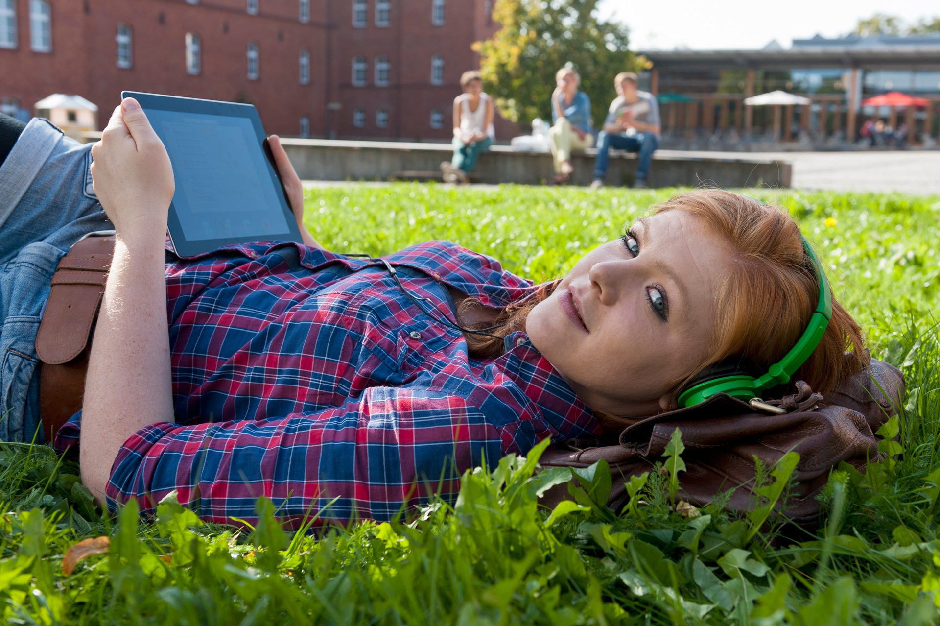 Junge Frau liegt im Gras, hört Musik und liest auf dem Tablet.
