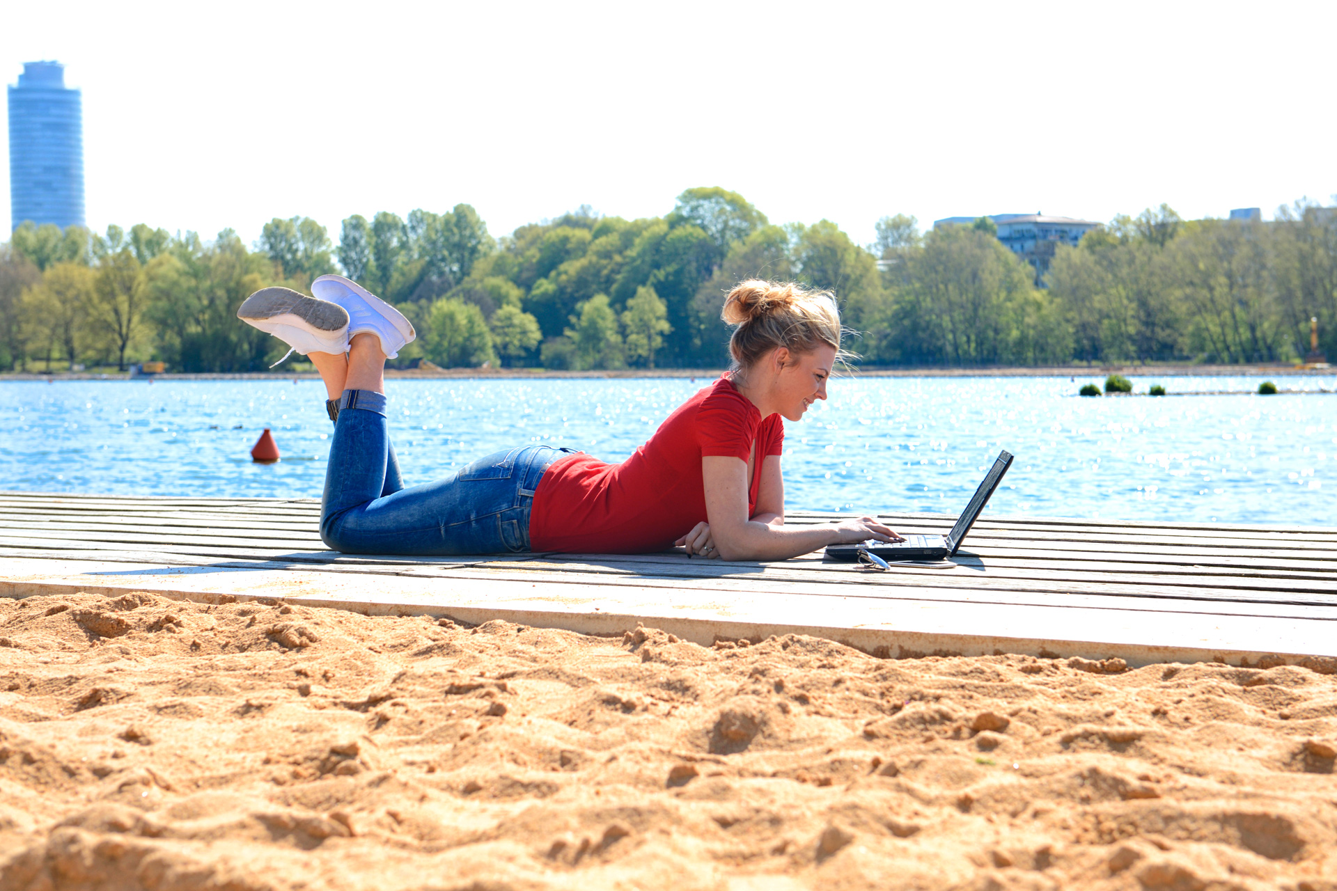 Eine Studentin liegt mit dem Laptop am Strand vor einem See.
